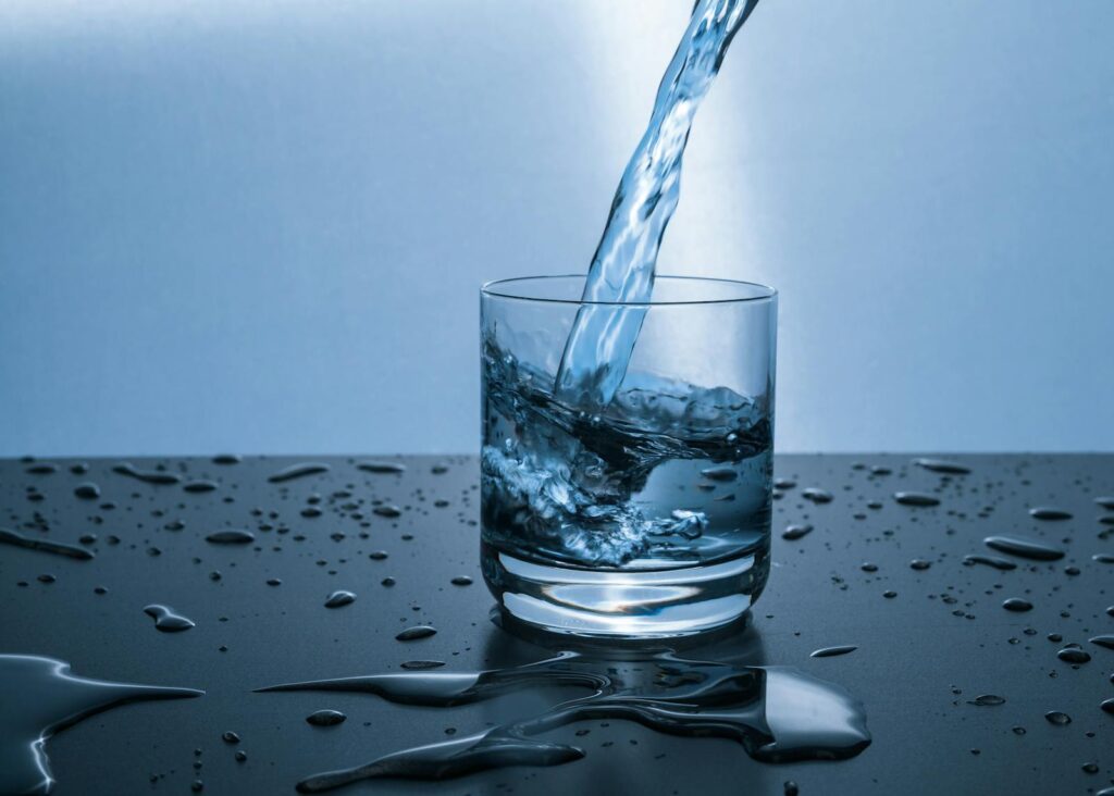 Agua fresca y cristalina vertida en un vaso sobre una superficie con gotas — el hábito sencillo de hidratación nocturna que le gana al "mouth taping" viral contra la boca seca al dormir.